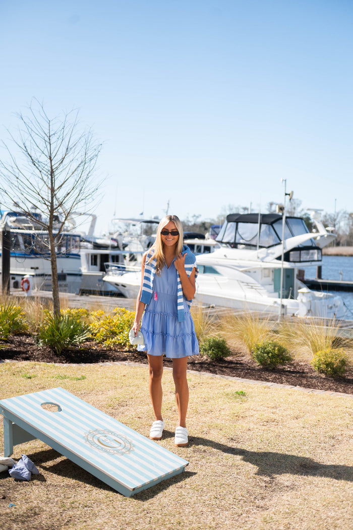 Model playing cornhole on a lawn in front of a marina in a mini length, sleeveless periwinkle dress. It is ruched at the waist and tiered. It has pink tassels hanging from the collar.