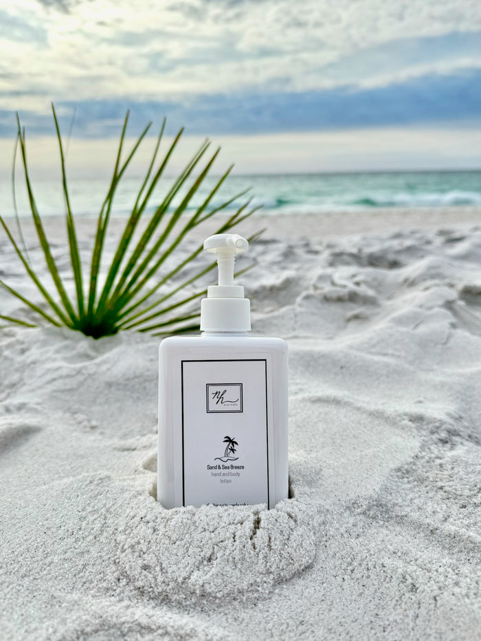 A bottle of Sand & Sea Breeze Lotions, standing in the sand on a beach with a green palm leaf and the ocean in the background.