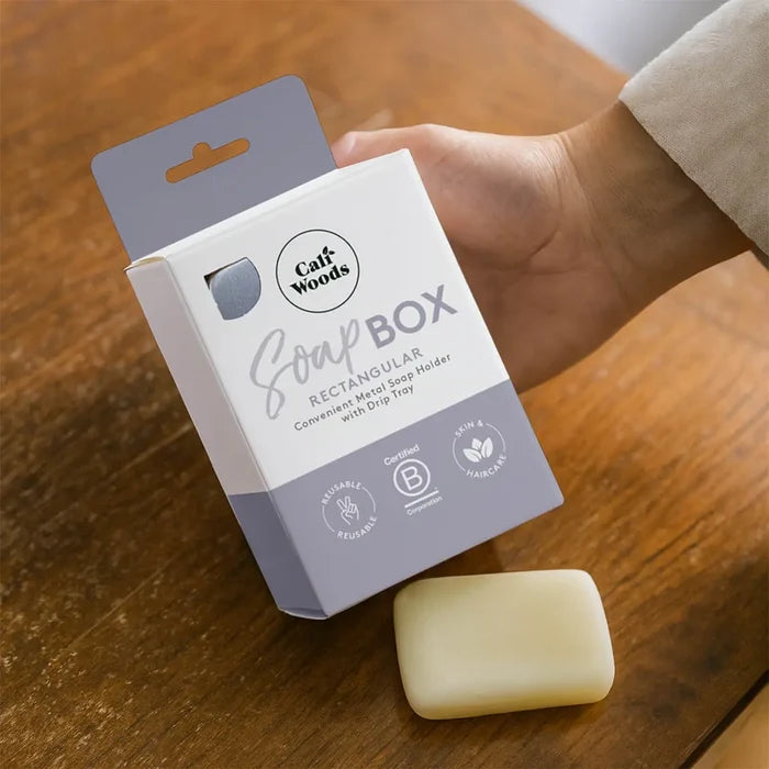 Hand holding a CaliWoods Soap Box packaging on a wooden surface with a bar of soap beside it.