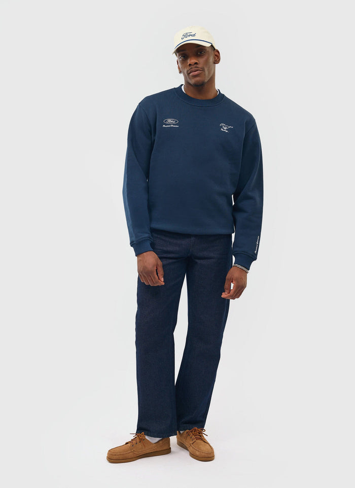 Model wears the navy embroidered Percival x Ford sweatshirt with dark jeans, brown shoes and a white Ford cap.