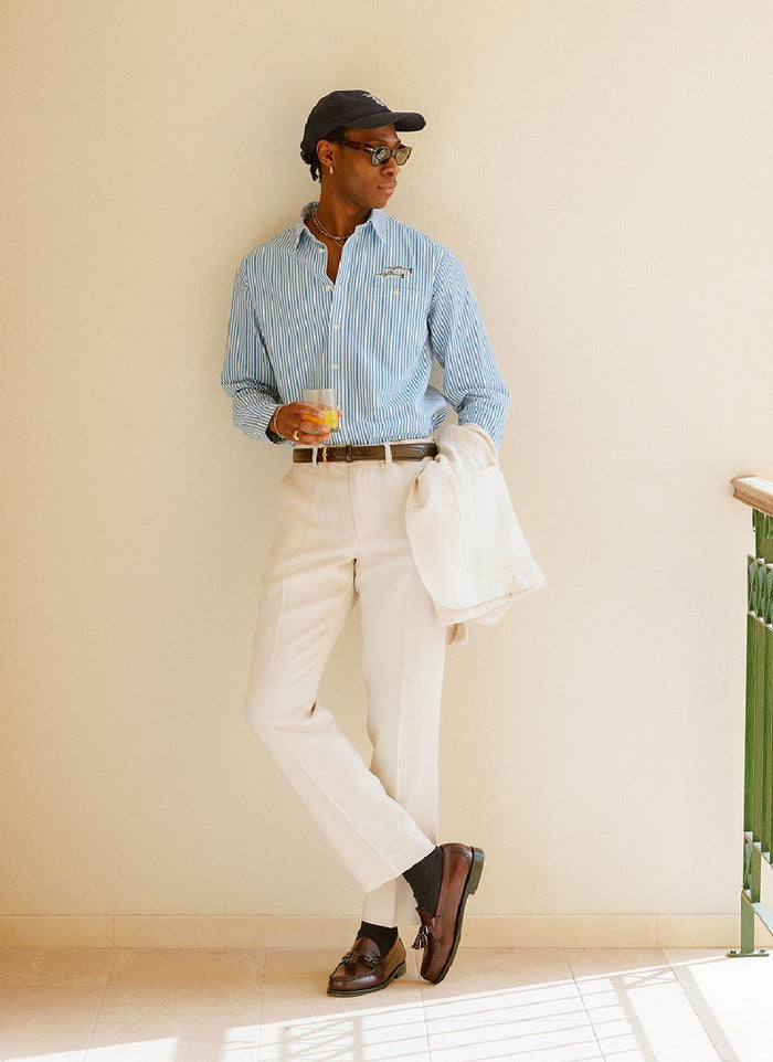 A model wears Percival's cream tailored linen trousers with a striped shirt, loafers, and matching blazer.
