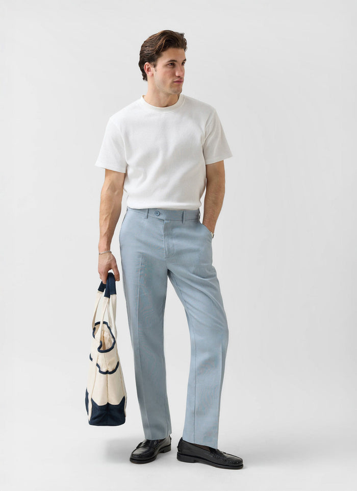 Model wears Percival Menswear light blue tailored linen trousers with a white t-shirt and black loafers, holding a tote bag.