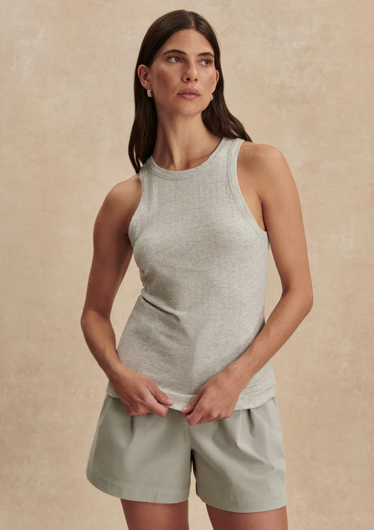 A woman modeling a light heather gray ribbed tank top and sage green shorts against a textured beige studio backdrop.
