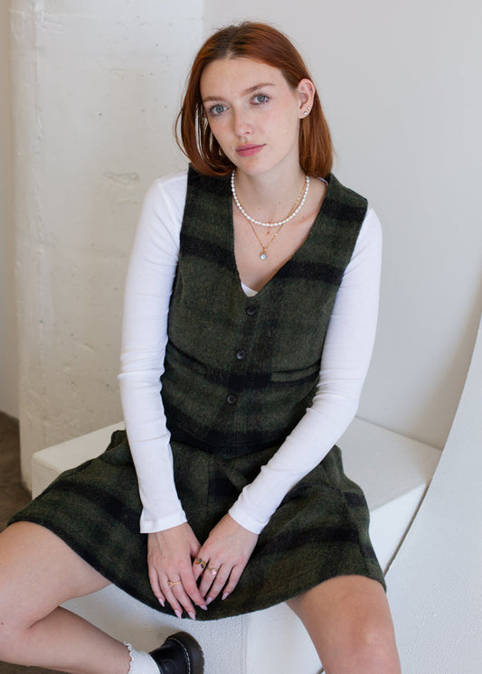 Woman wearing a green vest over a white long-sleeve shirt, sitting on a white chair.